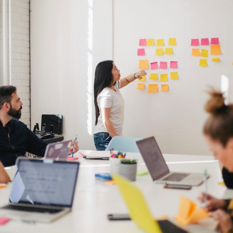 ingénierie de formation Une femme présente des notes autocollantes sur un mur, des personnes assises écoutent.